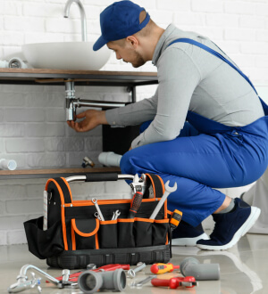 Plumber in blue overalls and cap fixing a pipe under a sink with a toolbox and various plumbing tools in the foreground.