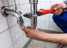 Person using a red pipe wrench to tighten plumbing under a sink with water dripping from the pipes.