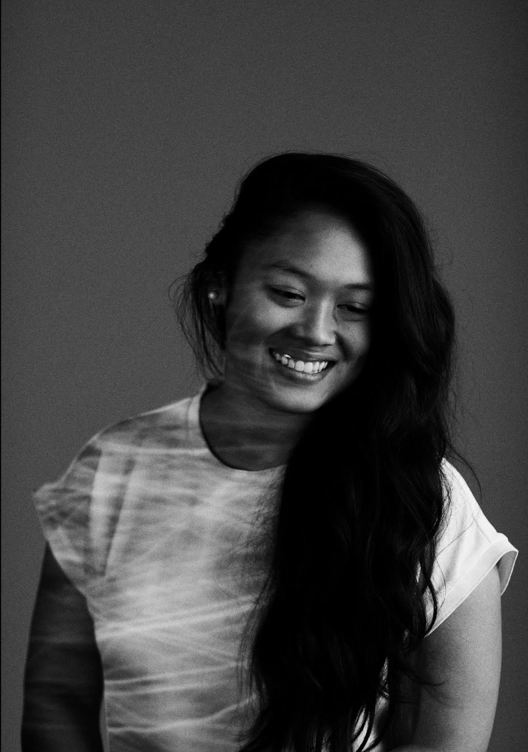 Black and white portrait of a smiling woman with long dark hair wearing a light-colored T-shirt against a plain background.