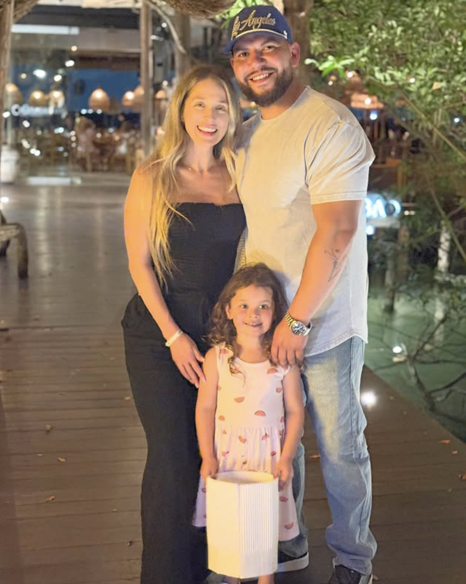 Smiling family of three posing on a wooden walkway at night, with the child holding a white lantern.