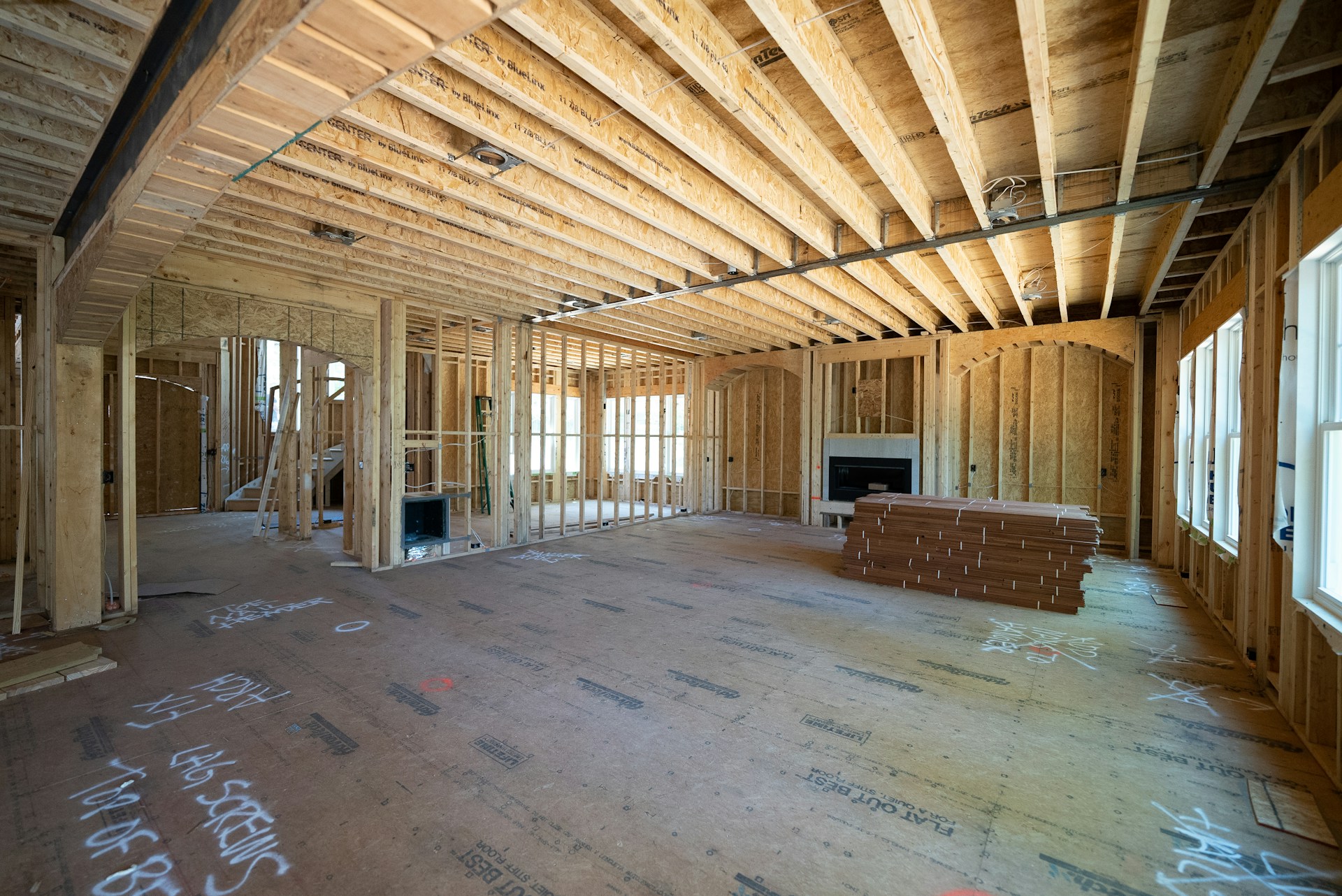 Interior of a house under construction showing exposed wooden framing, ceiling joists, and a partially built fireplace with stacked planks on the floor.