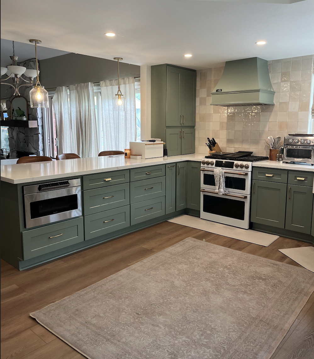 Modern kitchen with sage green cabinets, white countertops, stainless steel oven, gas stove, and light wood flooring with a beige patterned rug.
