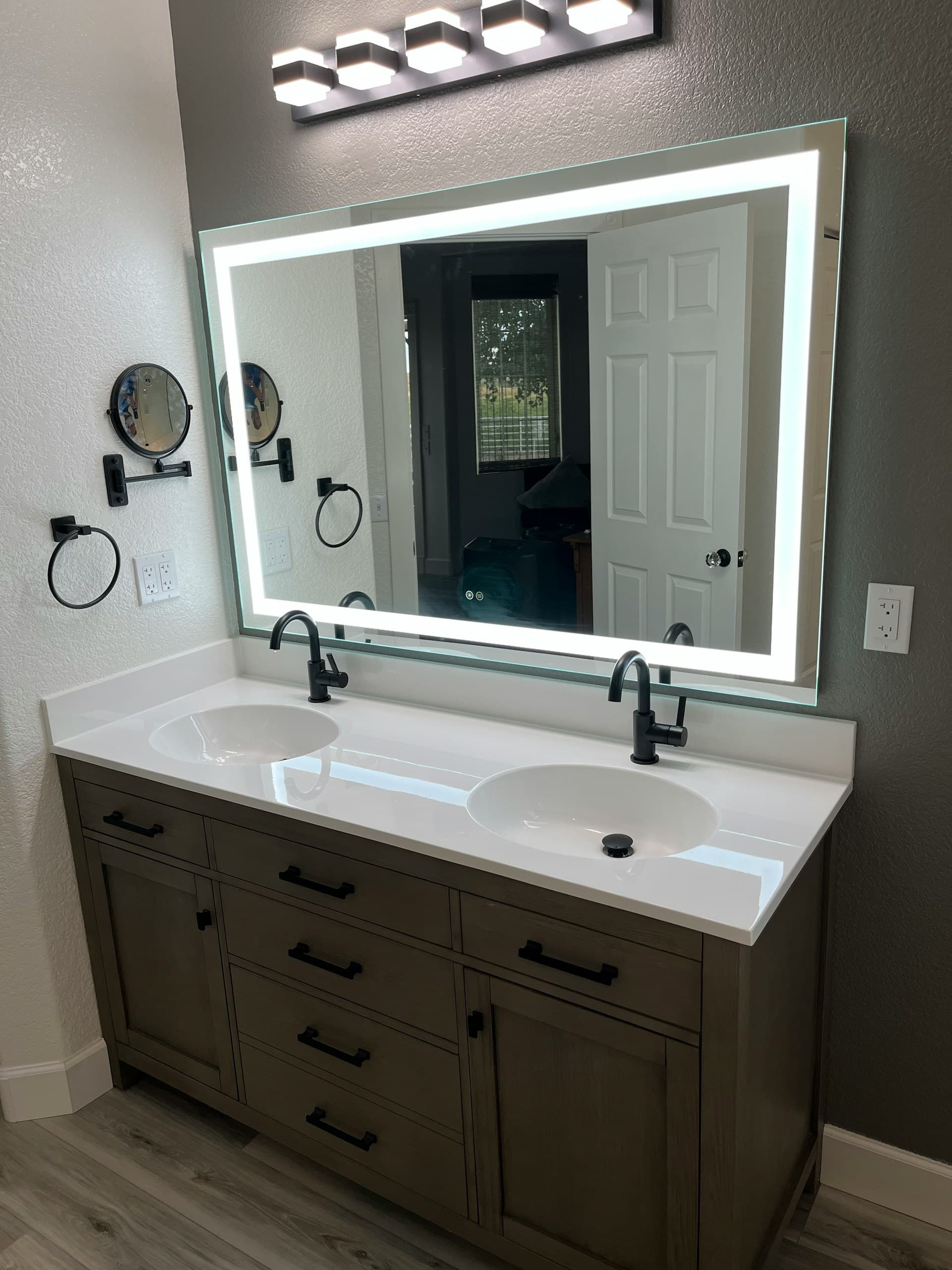 Modern bathroom with glass shower enclosure, bathtub with black fixtures, gray cabinetry, large mirror, and decorative dried flowers on the counter.