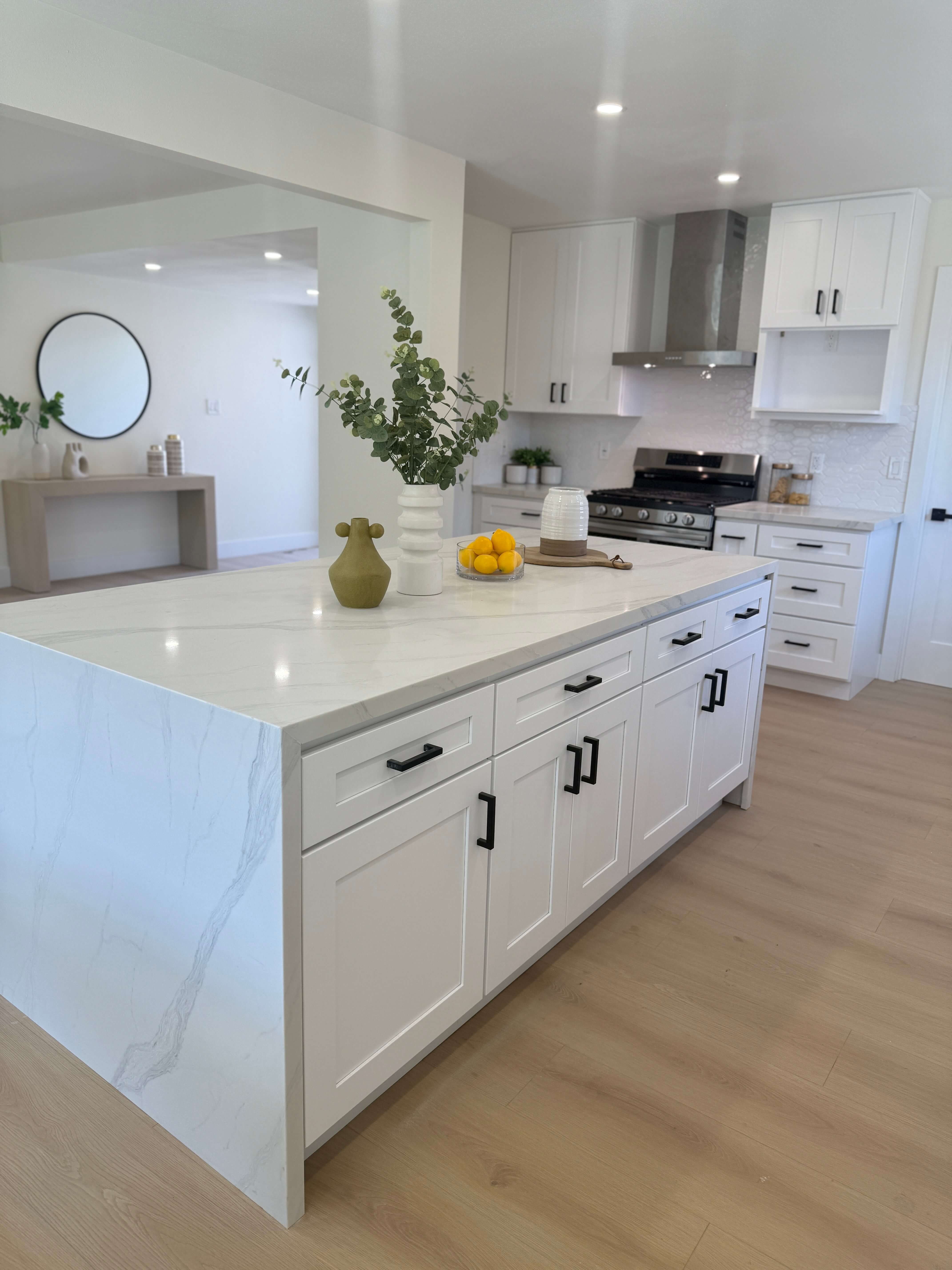 Modern bathroom with glass shower enclosure, bathtub with black fixtures, gray cabinetry, large mirror, and decorative dried flowers on the counter.