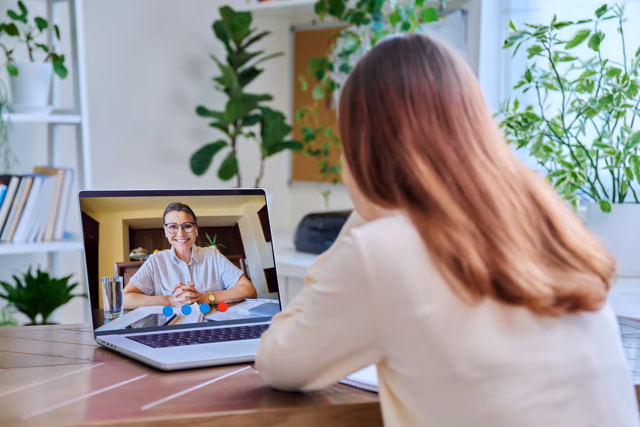 Woman having a virtual meeting via laptop with another woman smiling on the screen, surrounded by plants in a home office setting.