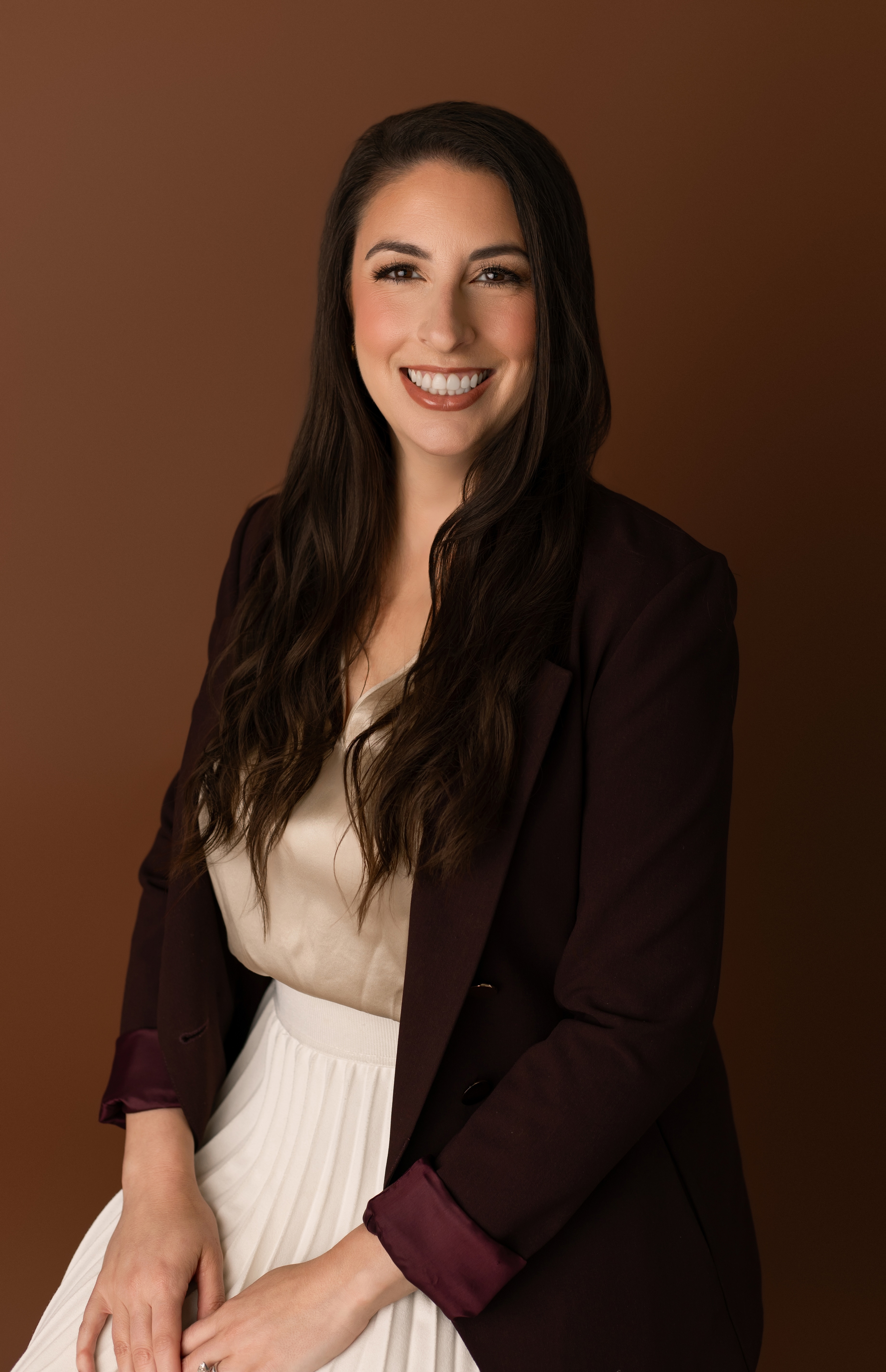 Smiling woman with long dark hair wearing a maroon blazer and cream blouse against brown backdrop.