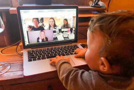 An adorable baby playing on a laptop with a video meeting for a creative marketing agency on the screen.