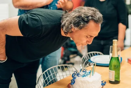 A man with dark hair blowing out candles on a birthday cake at a top creative marketing agency’s office. 