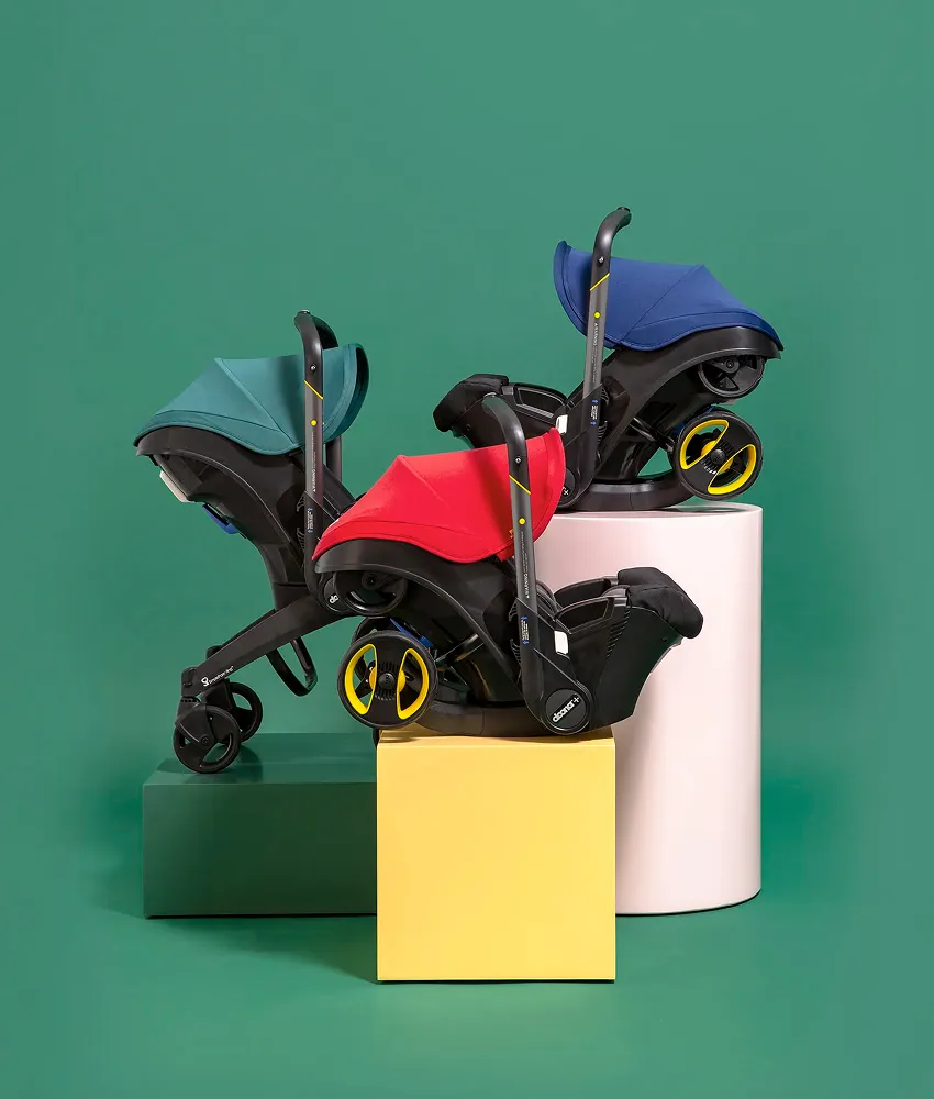 Three colorful Doona convertible car seat strollers on colorful podiums with a green background. 