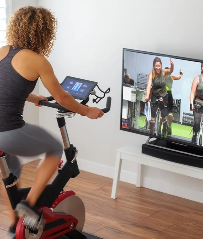 A Black woman riding a spin bike at home while watching a fitness video from Studio SWEAT onDemand.