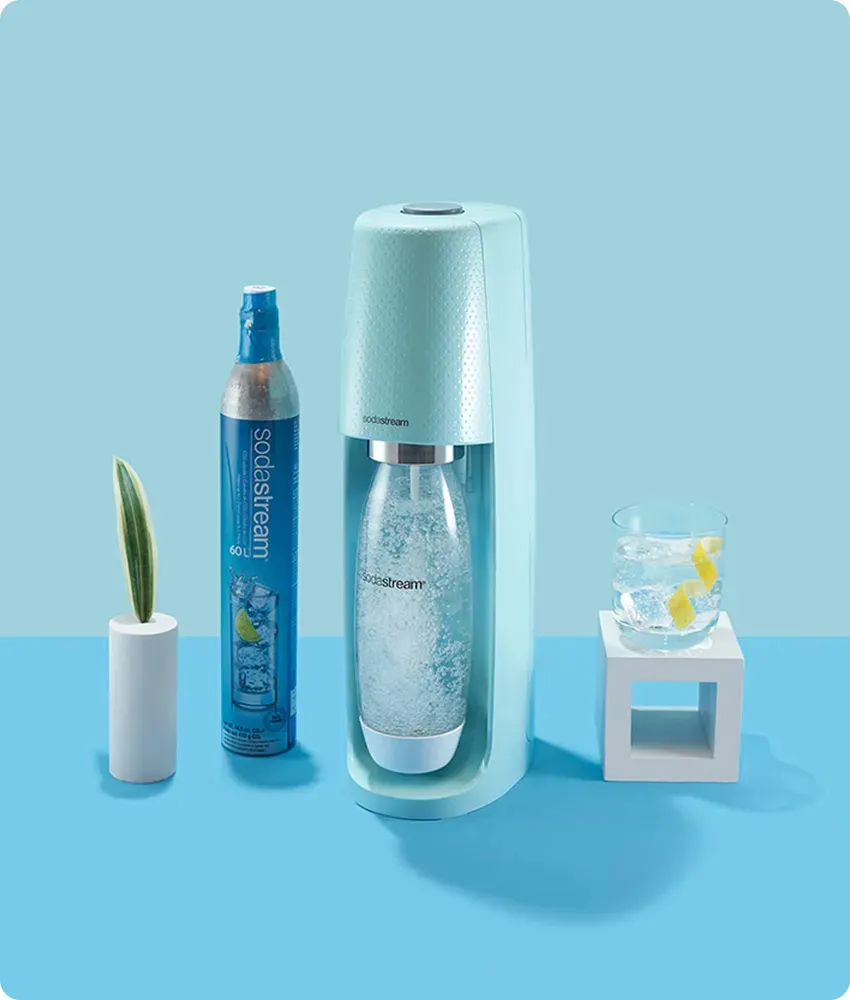 A blue sparkling water maker making a bottle of sparkling water next to a glass of water with a lemon wedge in front of a blue background. 
