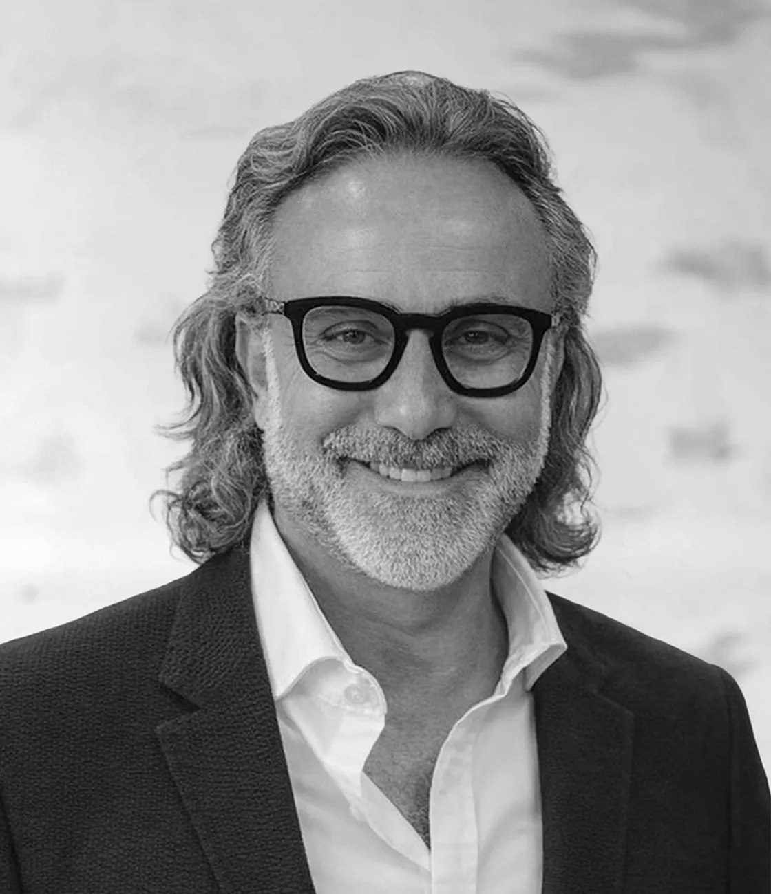 A handsome, ethnically ambiguous male marketing professional with long dark hair and a beard smiling, wearing thick black glasses and a suit.