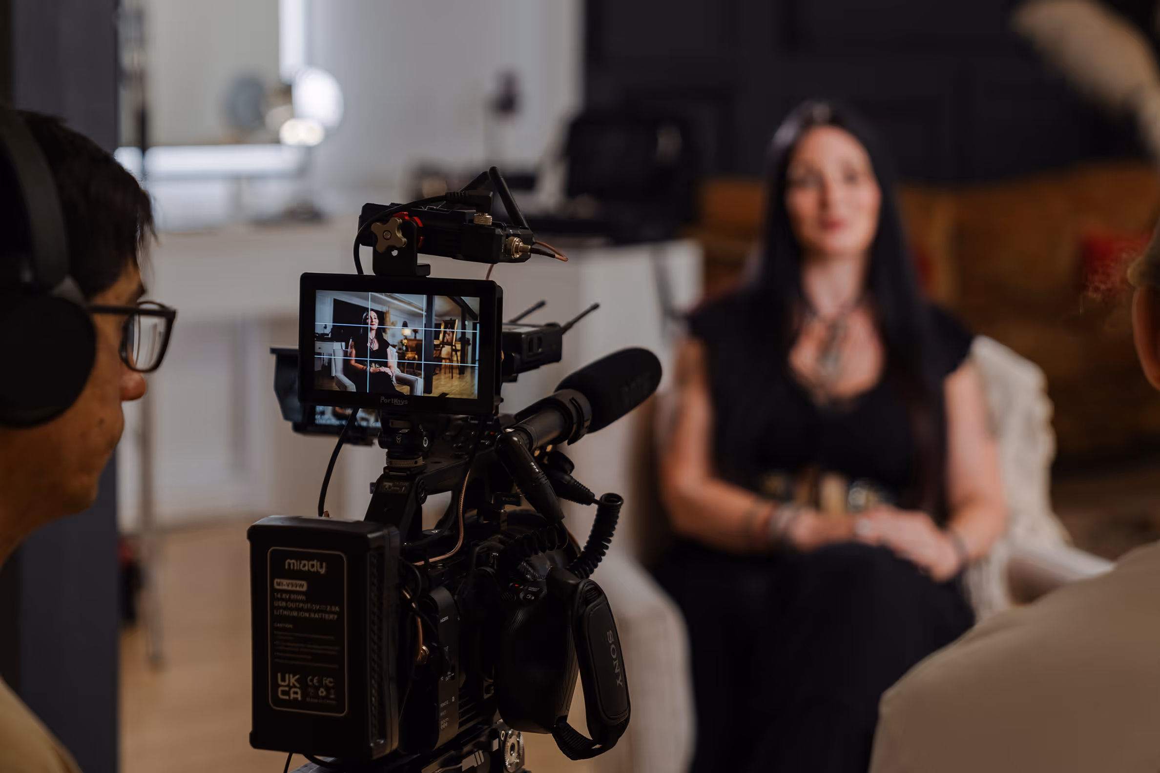 Video camera operator filming a woman sitting on a chair during an indoor interview or recording session.