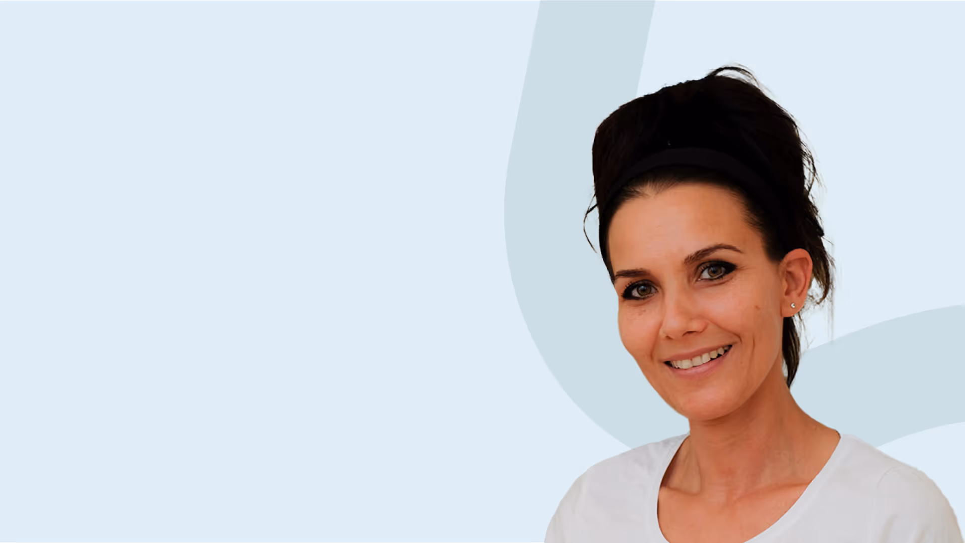 Lächelnde Frau mit dunklem Haar, das mit einem Stirnband zurückgehalten wird, vor hellblauem Hintergrund.