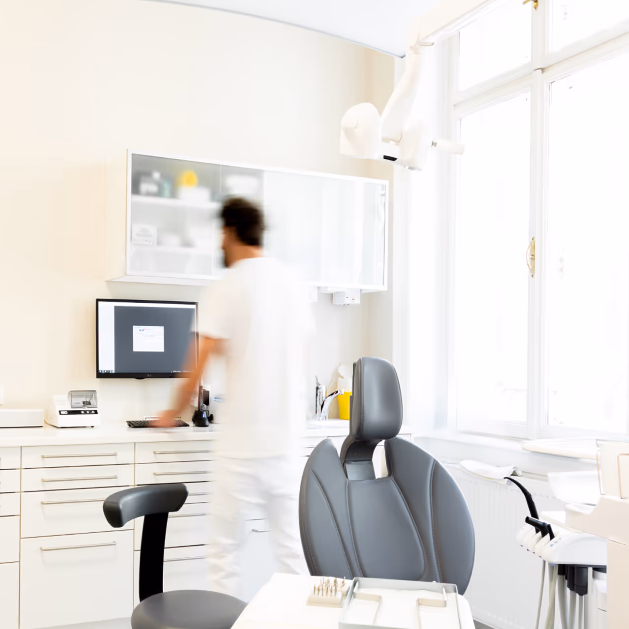 Zahnbehandlungsraum mit grauem Stuhl, Zahnärzteausrüstung und einem unscharfen Mann vor einem Computerbildschirm.