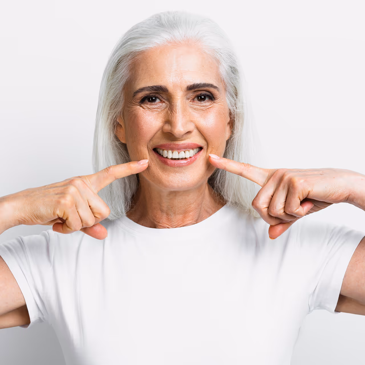 Ältere Frau mit weißen Haaren, die lächelt und mit beiden Zeigefingern auf ihr Lächeln zeigt.