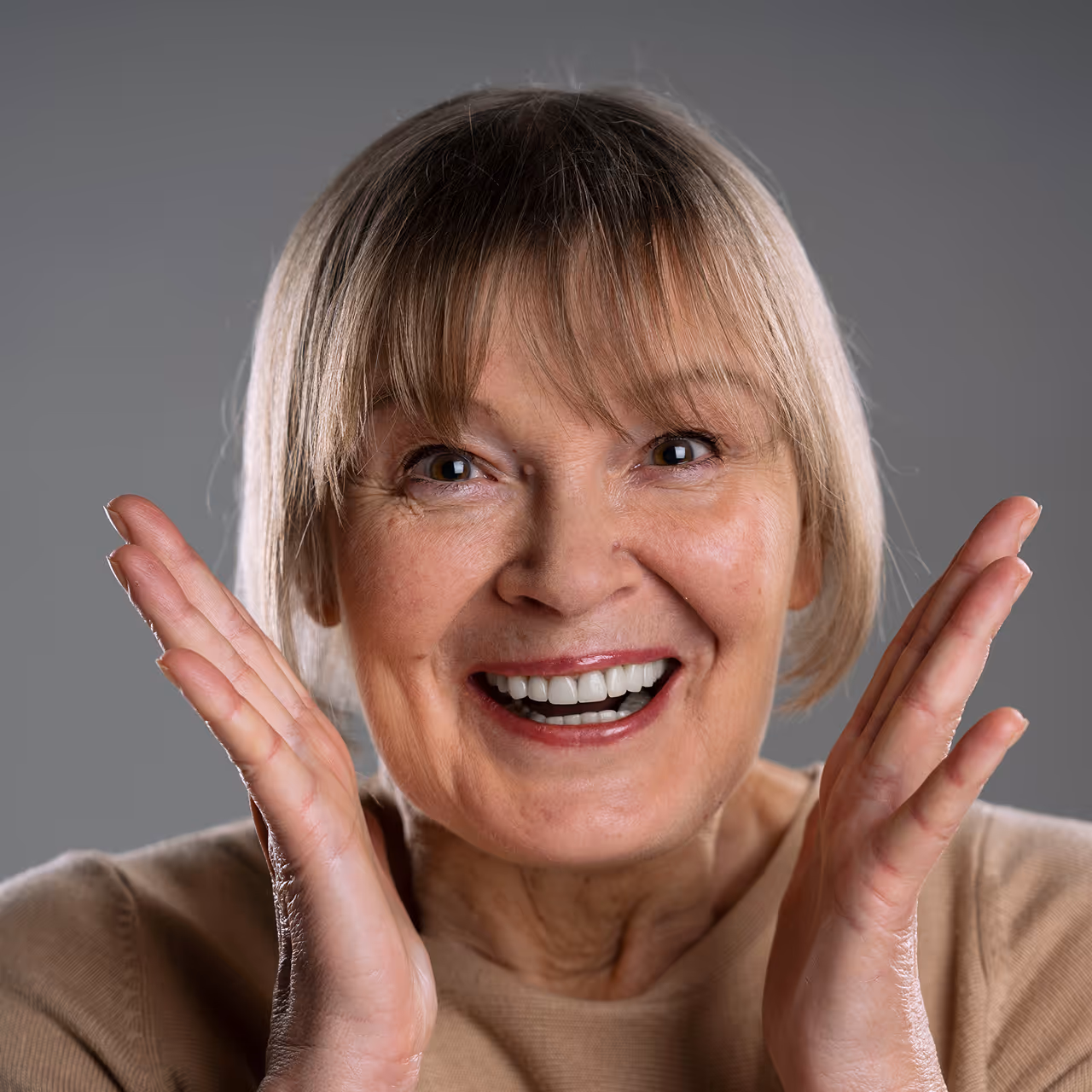 Ältere Frau mit blonden Haaren lächelt glücklich mit erhobenen Händen neben dem Gesicht.