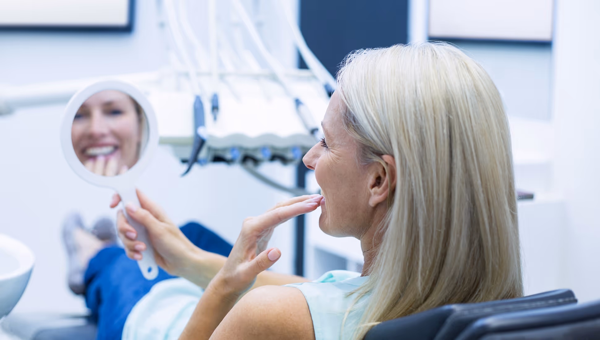 Frau mit blonden Haaren sitzt im Zahnarztstuhl und betrachtet lächelnd ihre Zähne im Handspiegel.