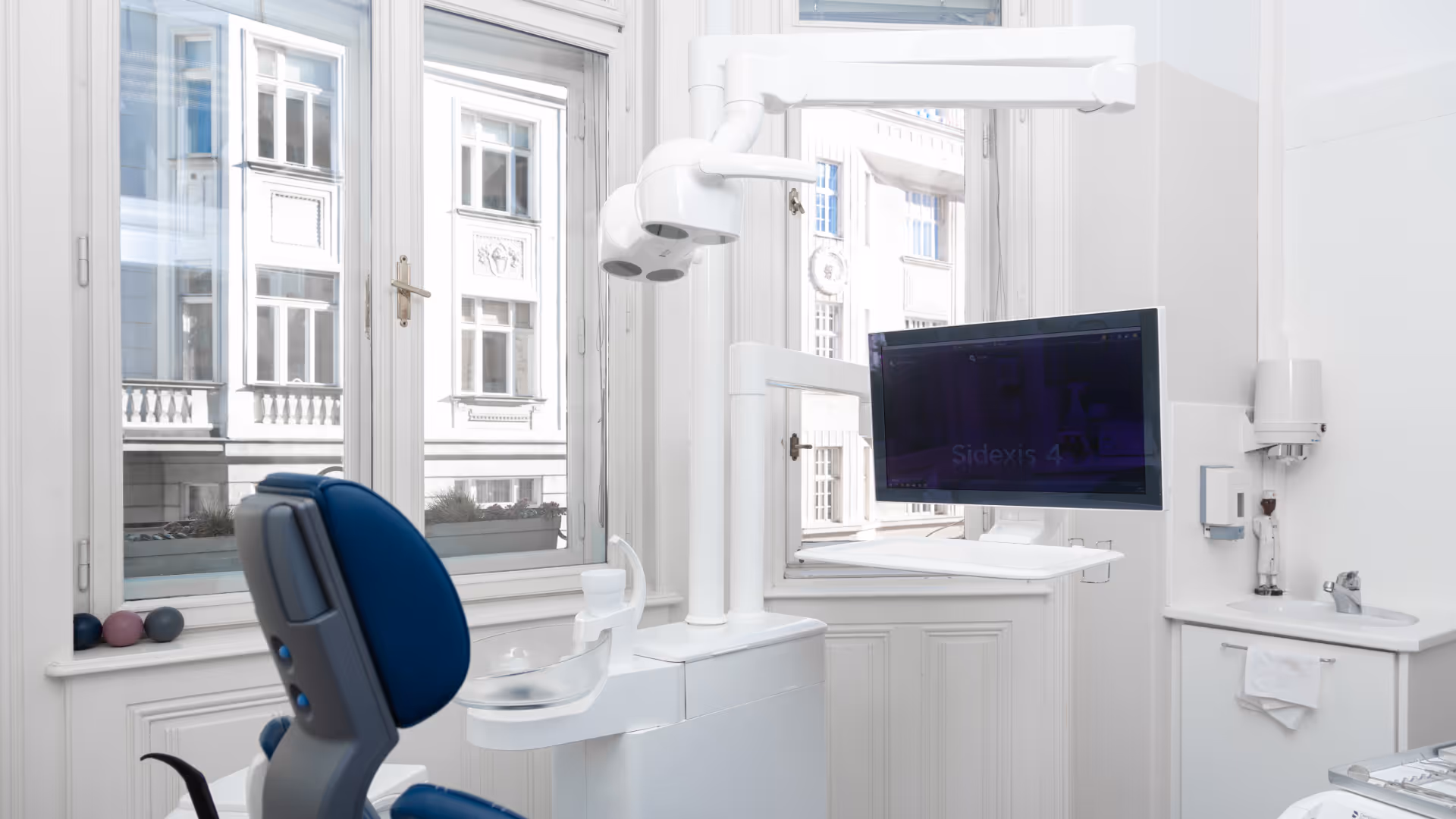 Hell gestalteter Zahnarztbehandlungsraum mit blauem Behandlungsstuhl, Monitor und großen Fenstern.