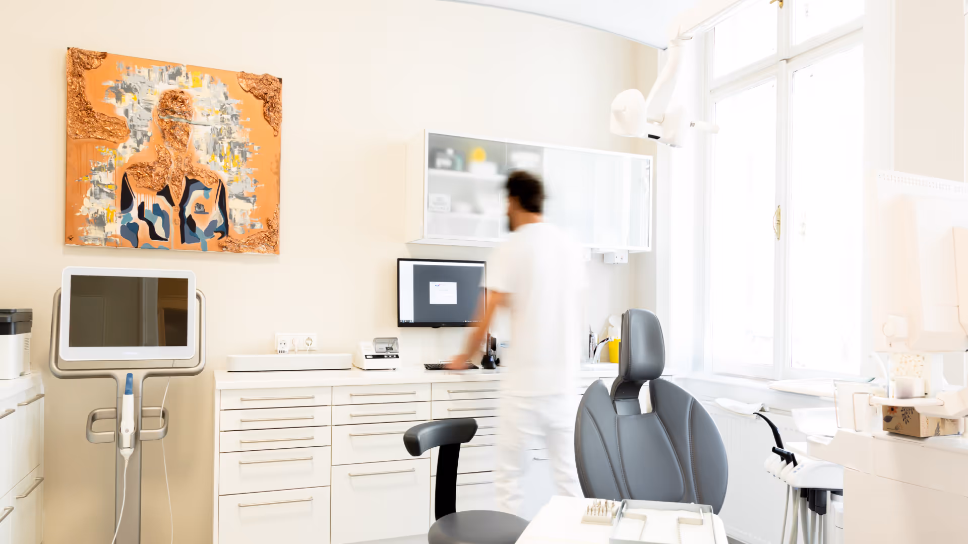 Helle Zahnarztpraxis mit grauem Behandlungsstuhl, Computerbildschirm und unscharfer Person in weißer Kleidung.