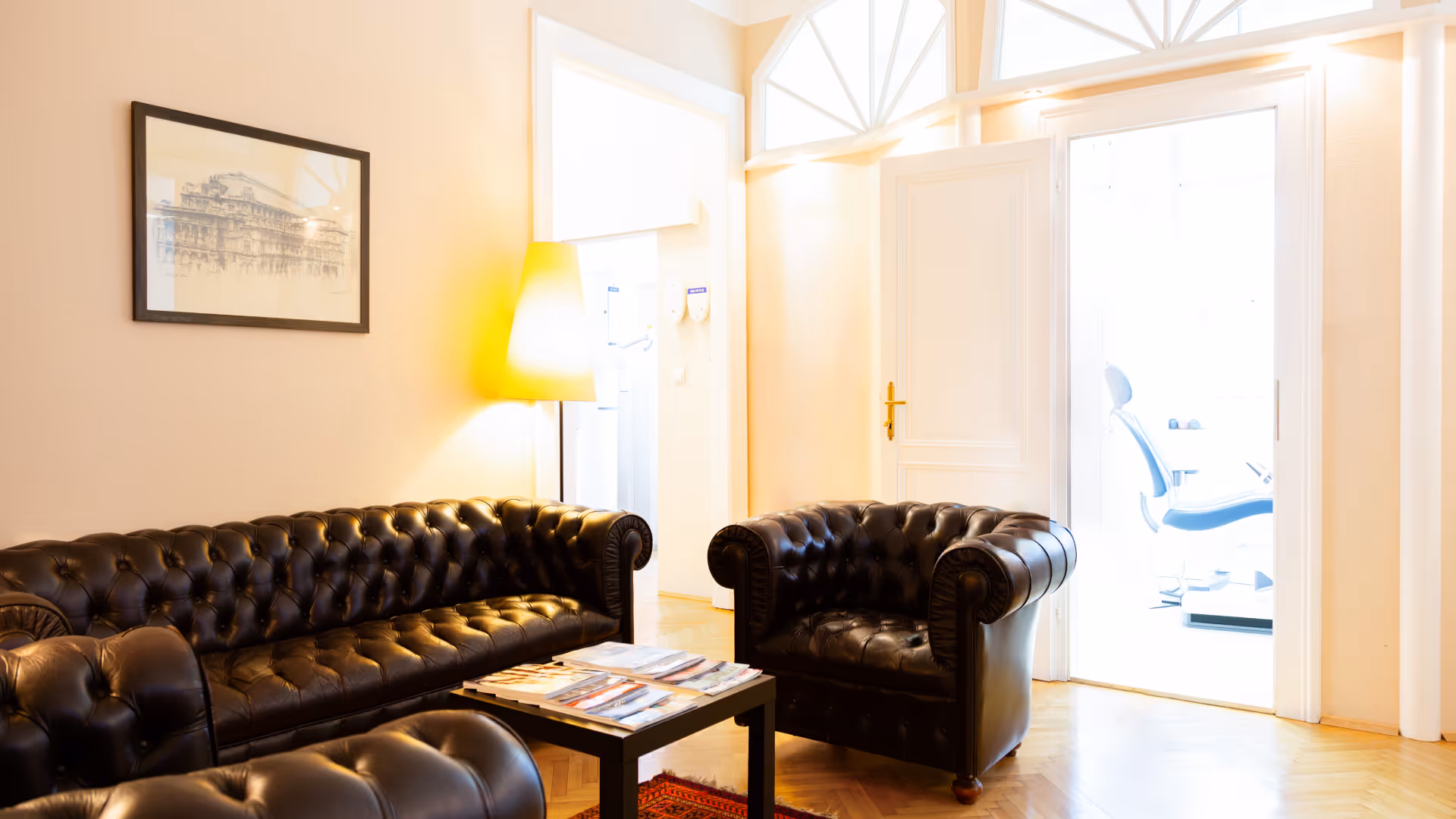 Helles Wartezimmer mit schwarzen Ledersofas, einem kleinen Tisch mit Zeitschriften und einer Stehlampe neben einem offenen Türrahmen.