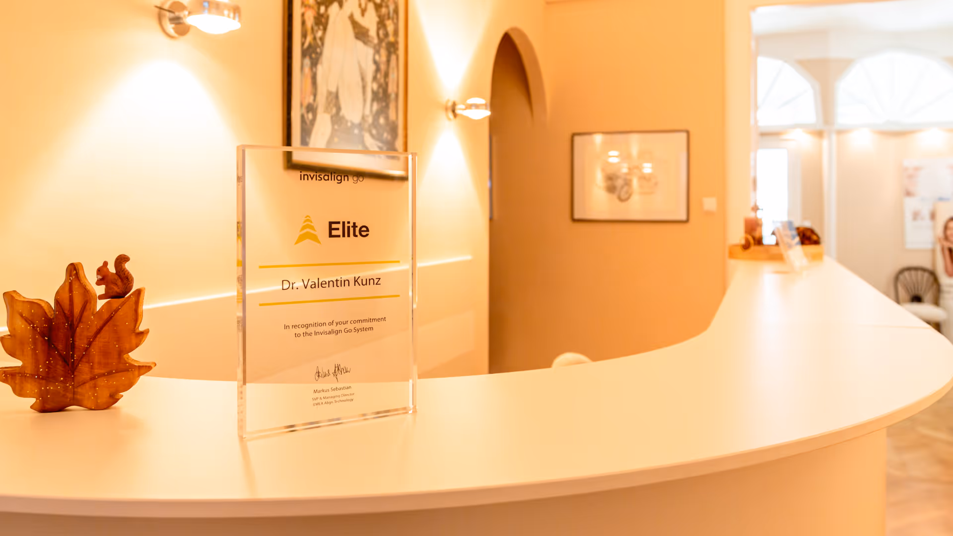 Empfangstresen mit einer Auszeichnung für Dr. Valentin Kunz als Invisalign Go Elite, neben einer Dekofigur eines Ahornblatts mit einem Eichhörnchen.
