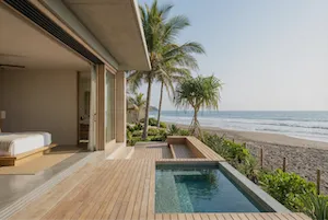 Modern beachfront villa with wooden deck, small pool, and sliding glass doors opening to a bedroom, overlooking the ocean and palm trees Proof El Salvador.