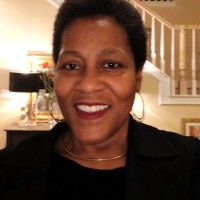 Smiling woman with short curly hair, wearing gold hoop earrings and a black top, standing indoors with stairs in the background.