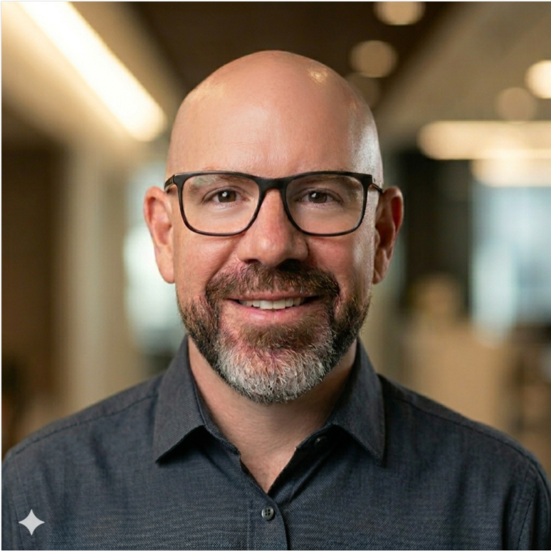 Smiling bald man with glasses and a beard wearing a dark button-up shirt in an office setting.