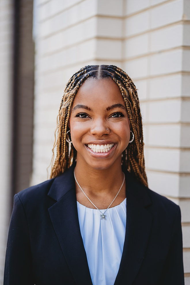 Smiling woman with braided hair wearing a black blazer, white shirt, and star necklace by a brick wall.