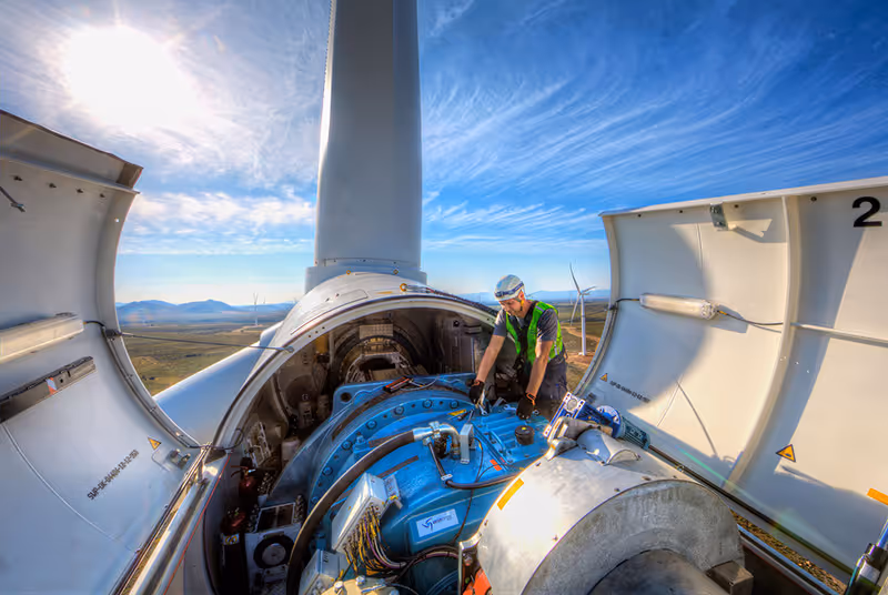 Technician in safety gear inspecting the inner machinery of a wind turbine under a partly cloudy blue sky.