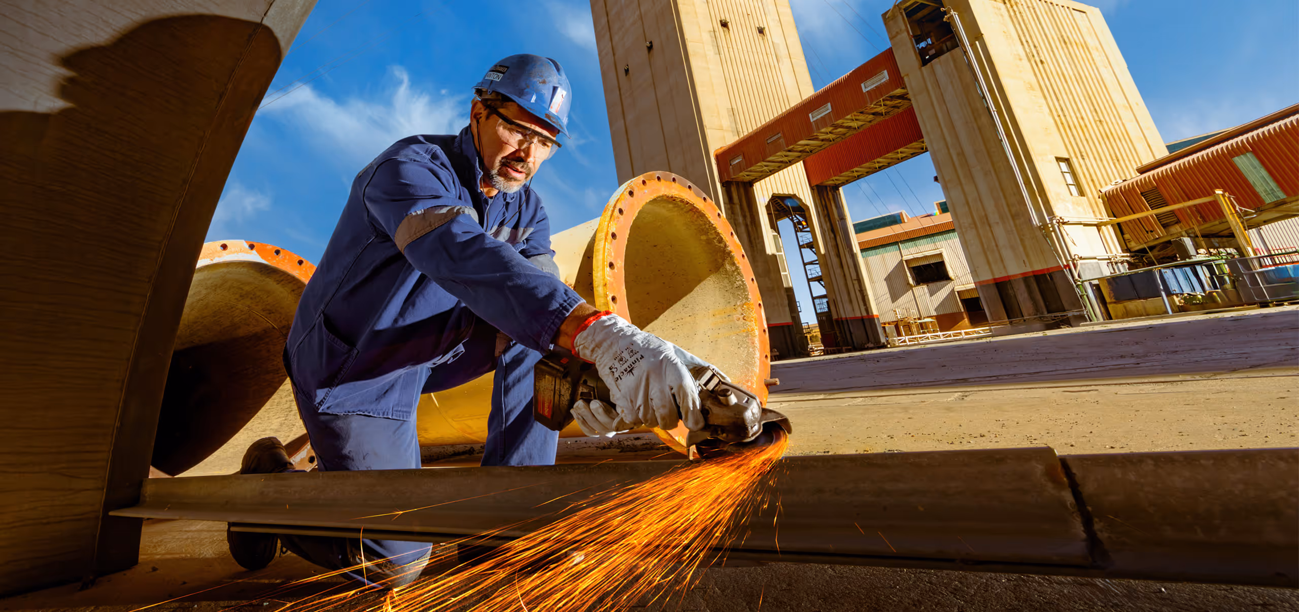 Worker wearing protective gloves and helmet using an angle grinder that sparks on a metal beam at a mine.