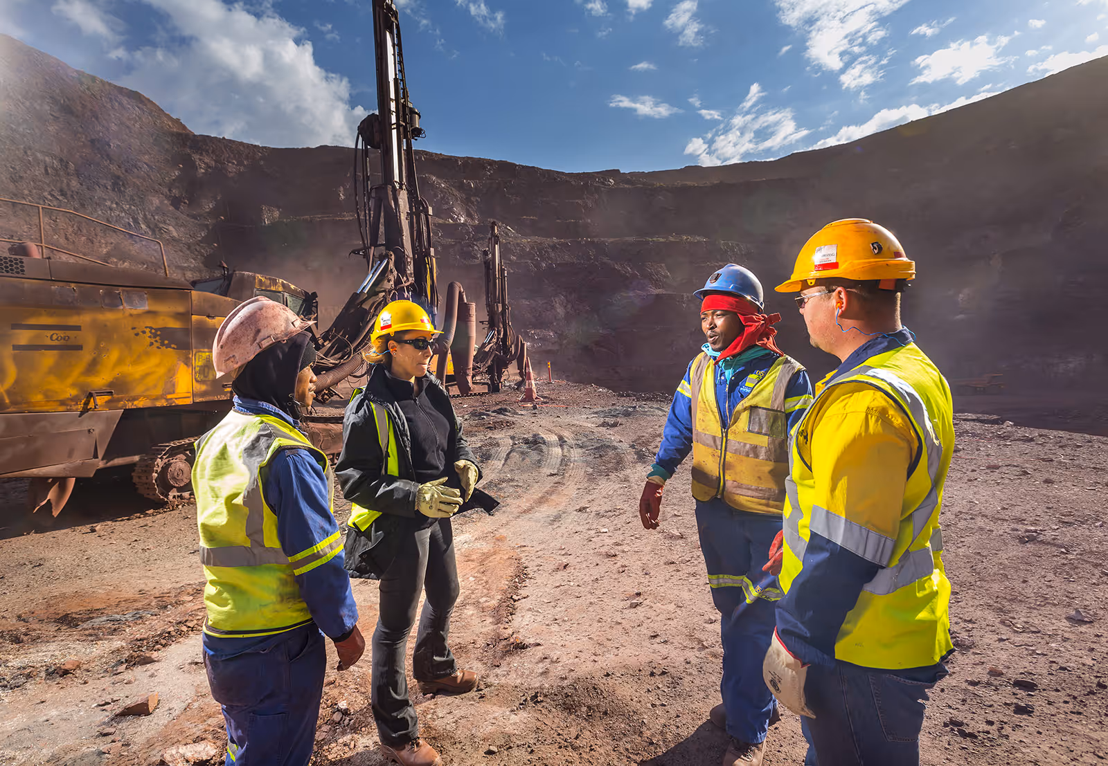 Four miners in safety gear having a discussion at an open-pit mine with drilling equipment in the background under a blue sky.