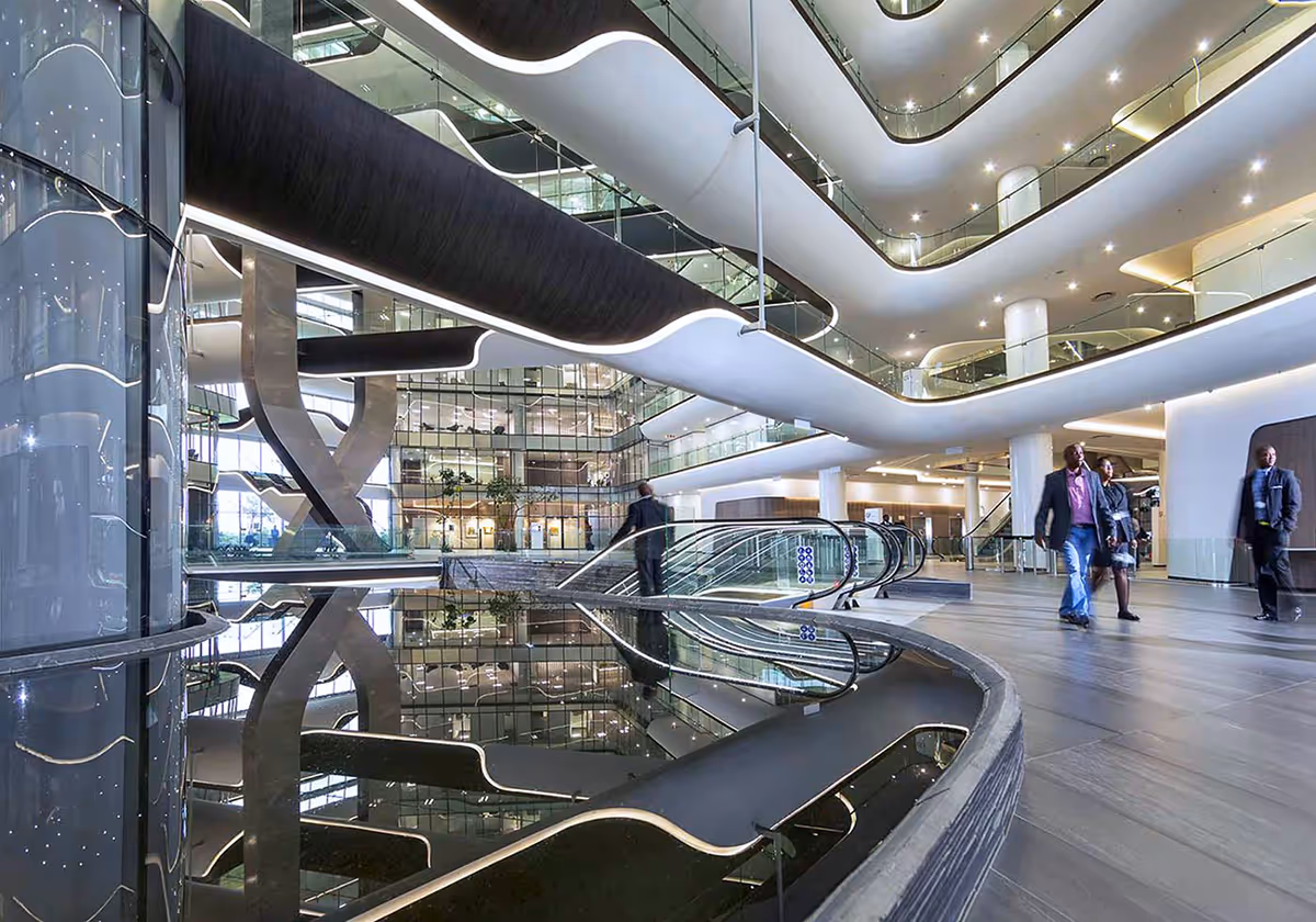 Architectural photography of modern corporate lobby with sleek curved balconies, escalators, reflective water feature, and people walking.