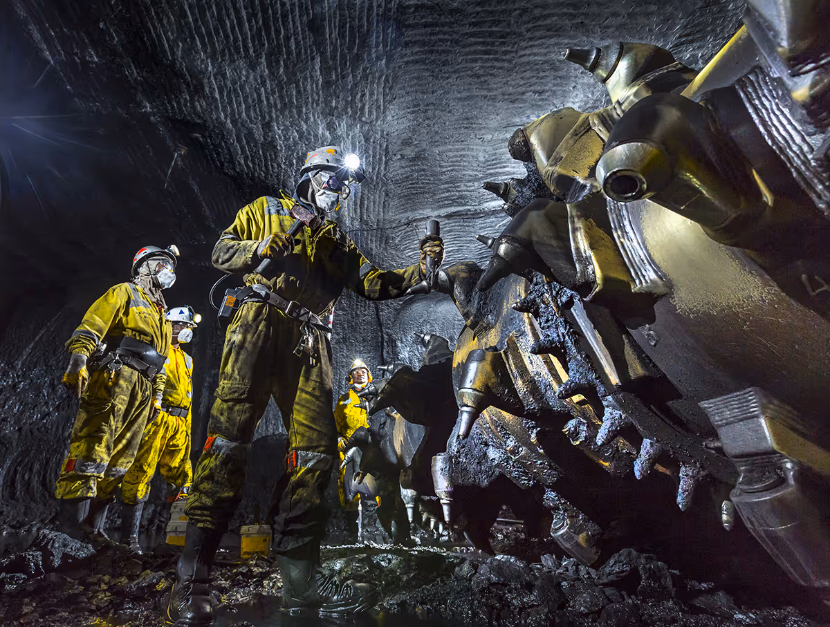 Four miners in yellow PPE with helmets and headlamps inspecting heavy mining machinery underground.