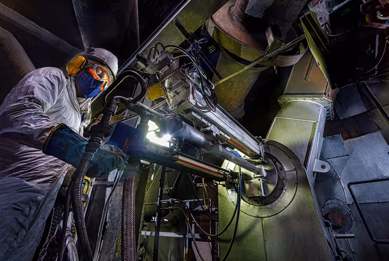 Worker in protective gear operating industrial machinery with metal pipes and hoses in a factory setting.
