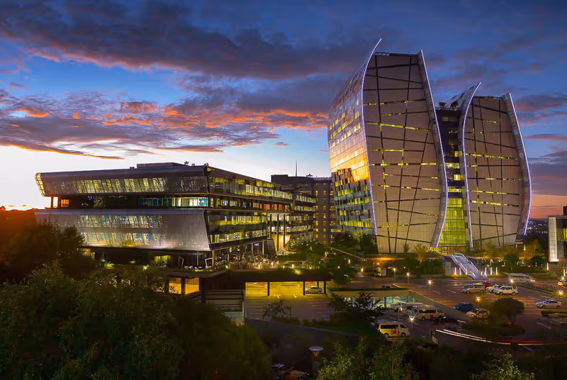 Modern office buildings with glass facades illuminated at dusk under a colorful cloudy sky.