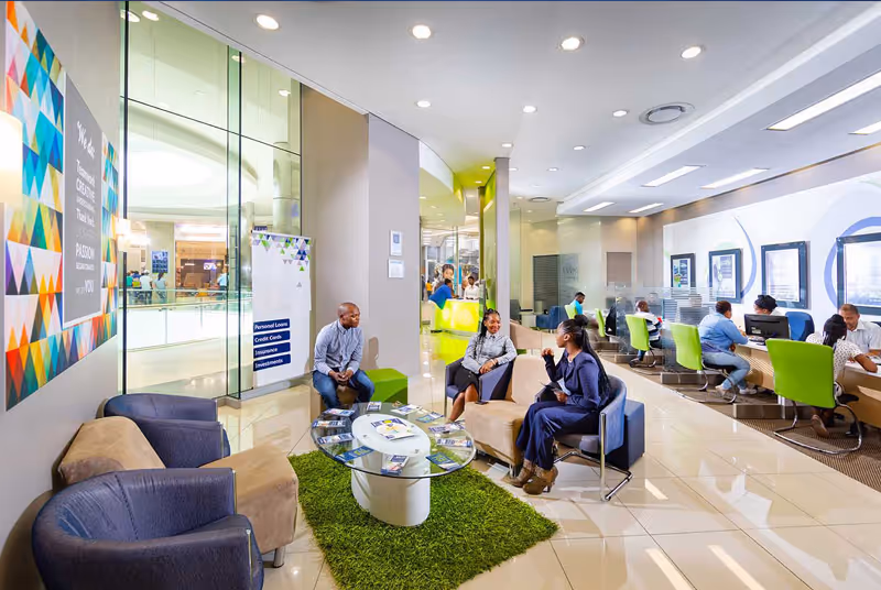 Bright modern office lounge area with people sitting on chairs and sofas around a glass coffee table on a green rug, and workers using computers in the background.