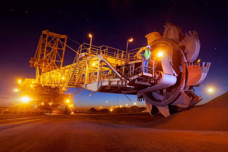 Large mining machine with a worker in safety gear standing on its platform, illuminated at night against a dark sky.