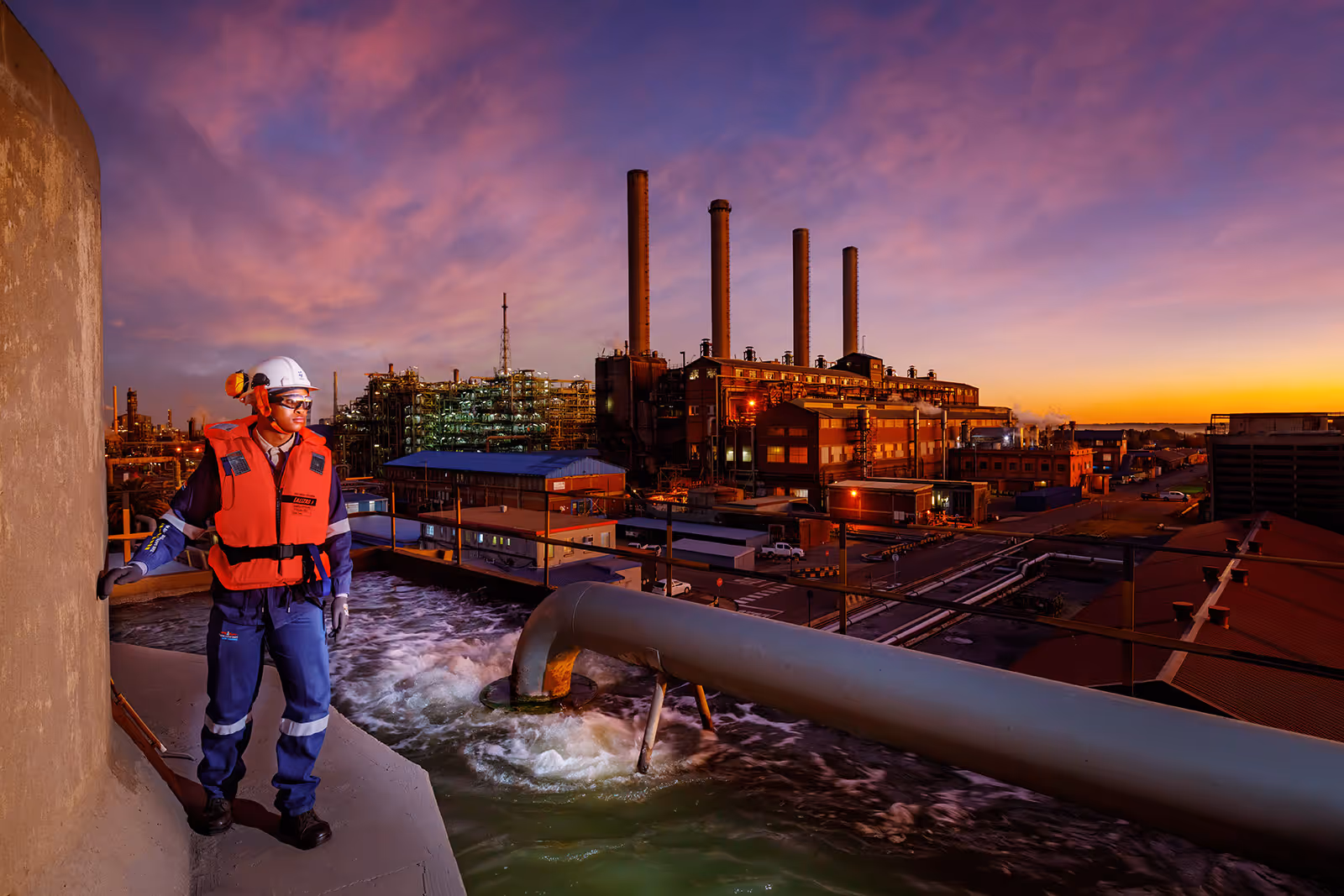 Worker in safety gear standing near industrial water outflow at a factory during sunset.