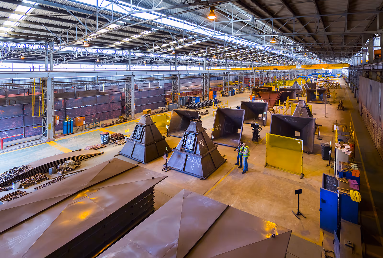 Large industrial warehouse with metal fabrications and two workers in high-visibility vests discussing near pyramid-shaped metal structures.