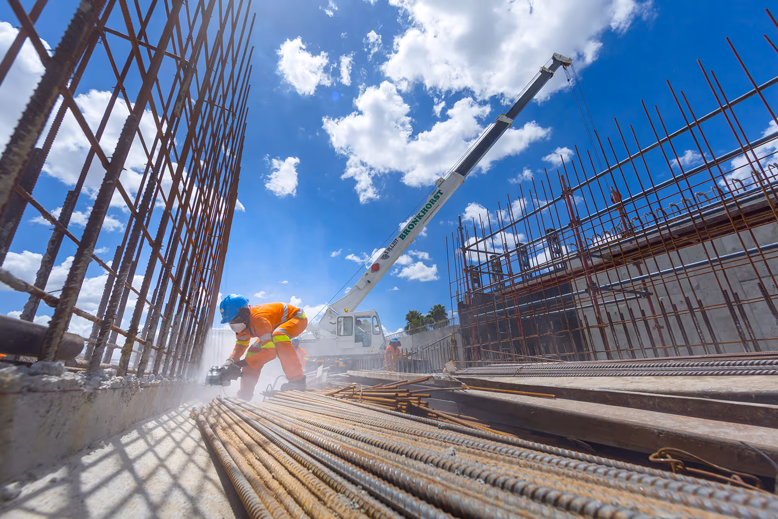 Construction worker in orange safety gear and blue helmet using a power tool on metal rods at a construction site with a crane and blue sky in the background.
