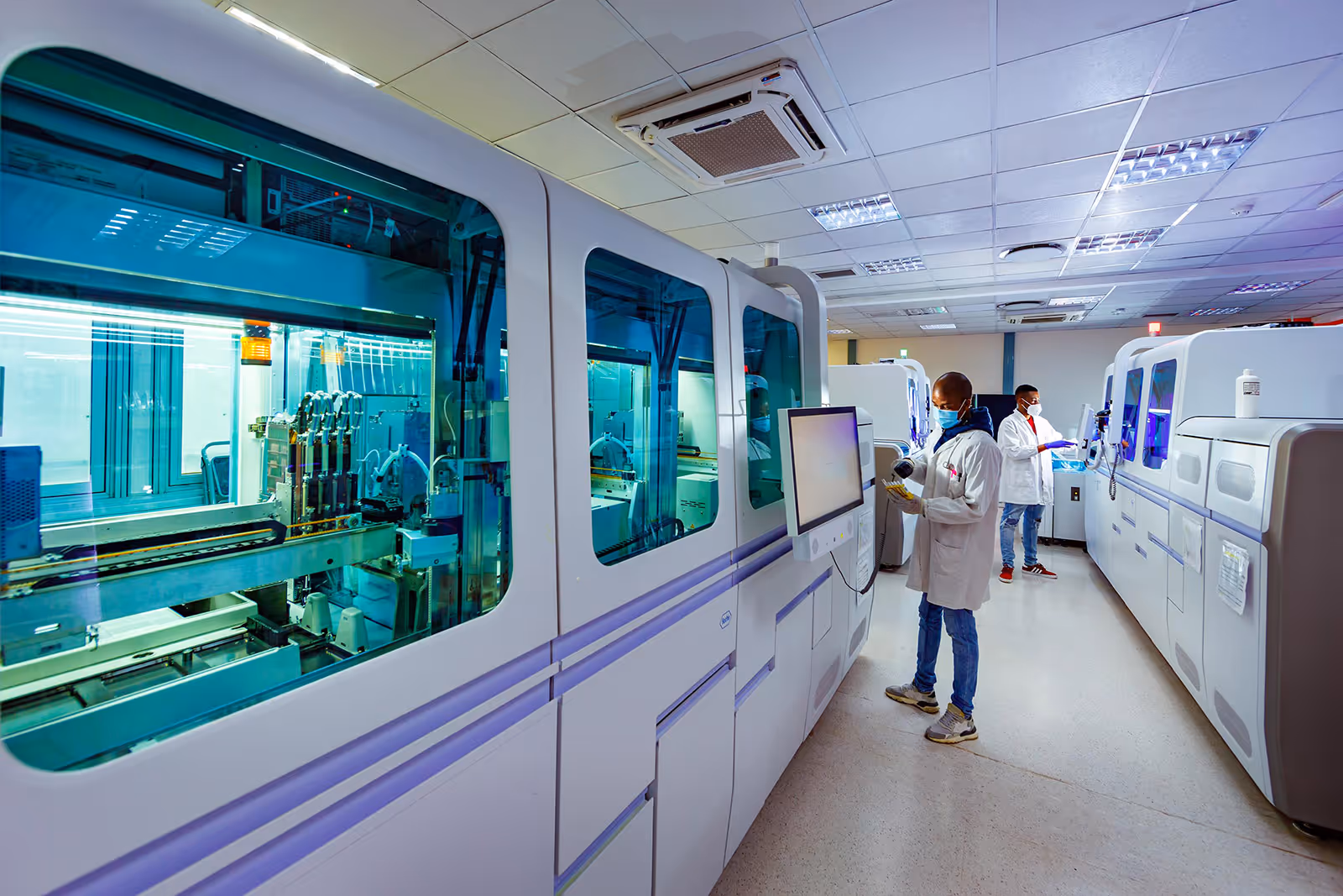 Two lab technicians wearing masks and white coats operating large automated laboratory machines in a clean clinical setting.