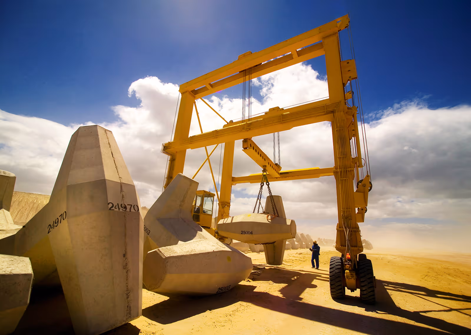 Yellow crane lifting a large concrete block with numbered tetrapods on sandy ground under a partly cloudy blue sky.