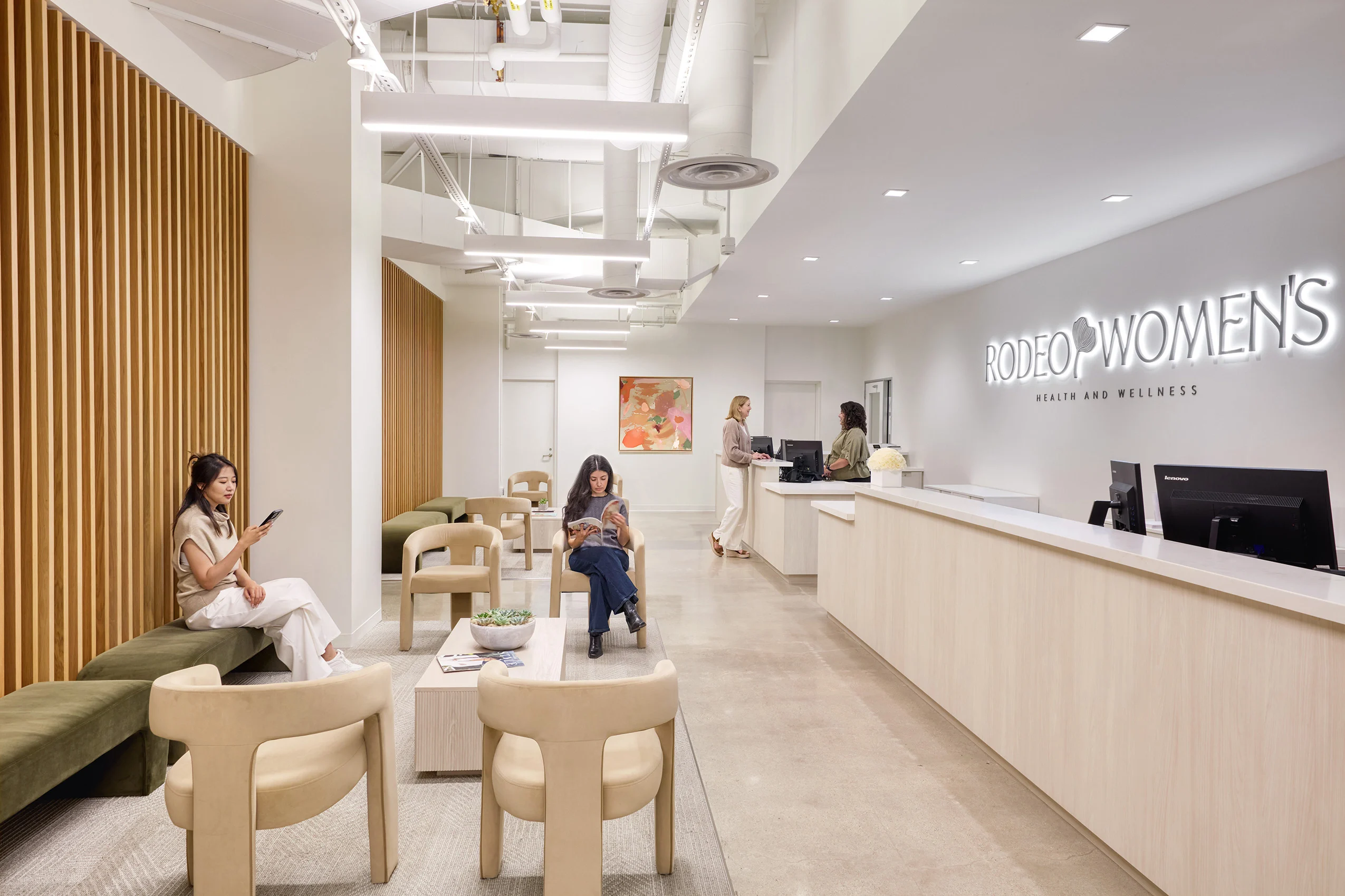 Modern health clinic waiting area with neutral tones. Three women sit with phones, and two stand at a reception desk under a "Rodeo Women's Health" sign.
