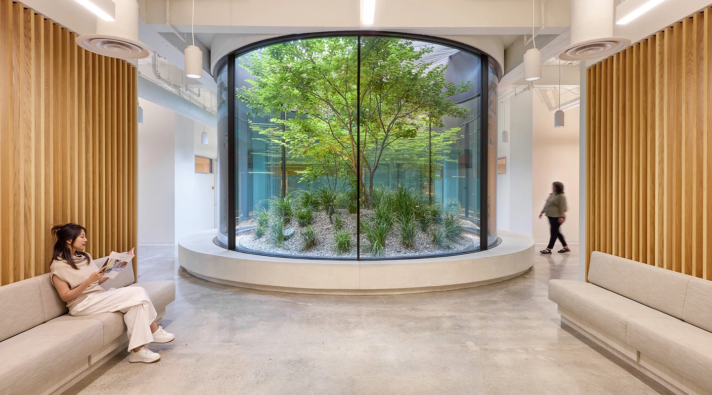 Modern lobby with minimalist design features a central circular glass planter with a tree and plants. A woman sits reading on a bench, conveying calmness.
