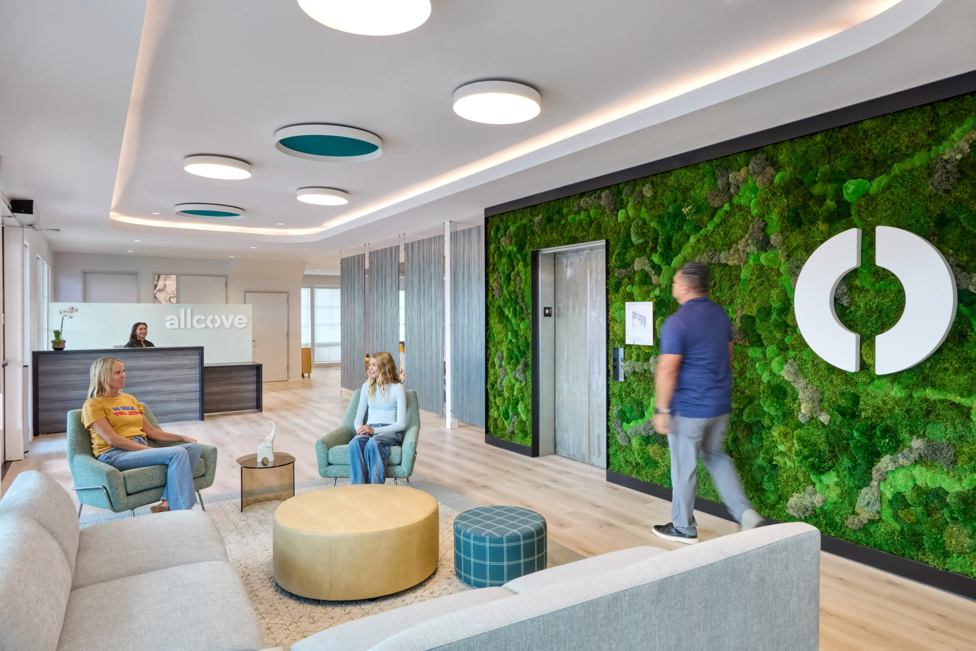 Modern office lobby with a green moss wall, sleek ceiling lights, and a reception desk. Two people sit on chairs, another walks past, creating a calm and professional atmosphere.