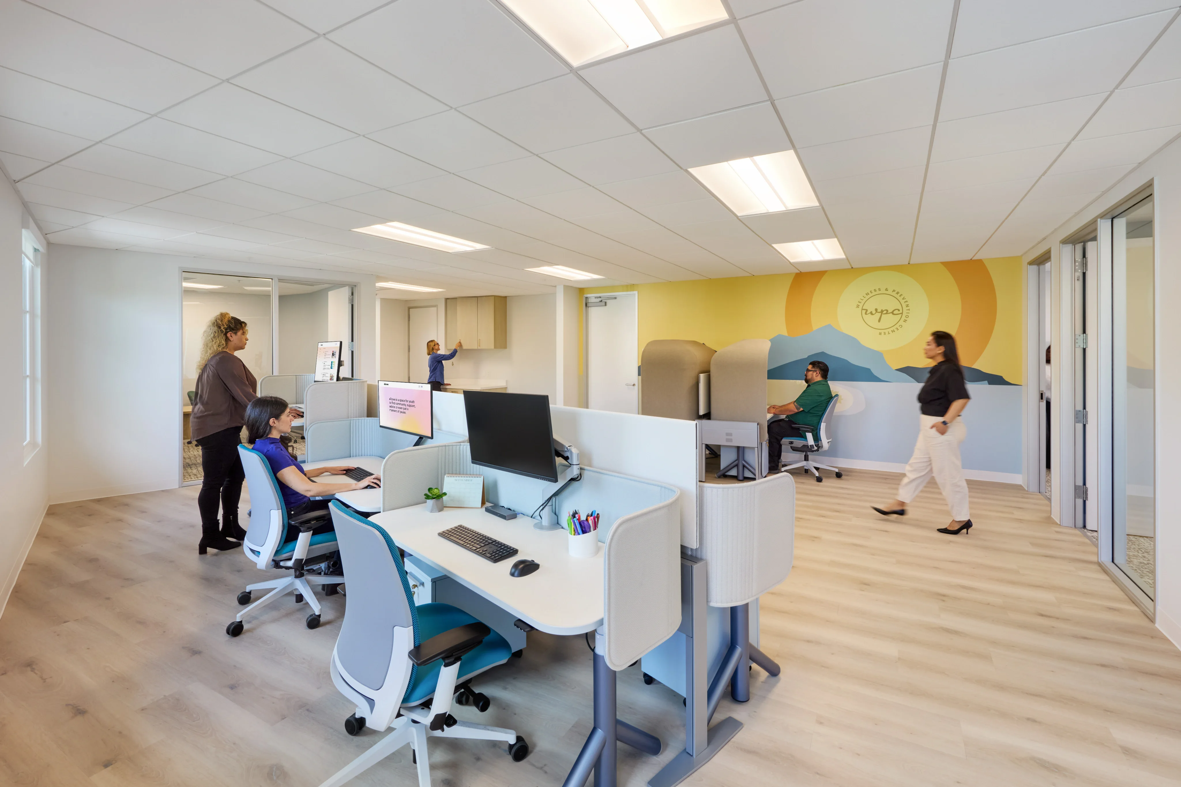 Modern office with bright lighting, people working at desks and walking. A calming mural on the wall depicts a sun and mountains, creating a serene atmosphere.