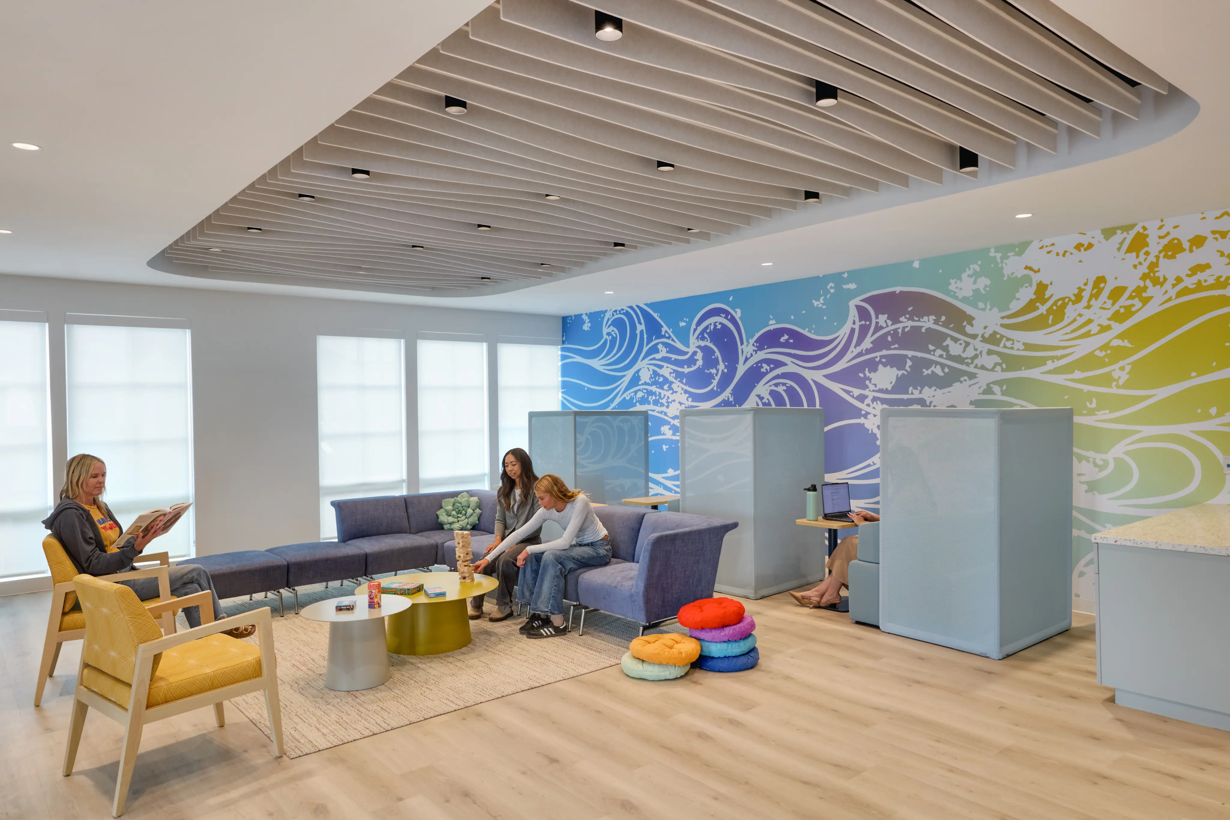 Modern lounge with a curved sofa, colorful wave mural, and cozy seating pods. Relaxed scene with people reading and chatting on a light wood floor.
