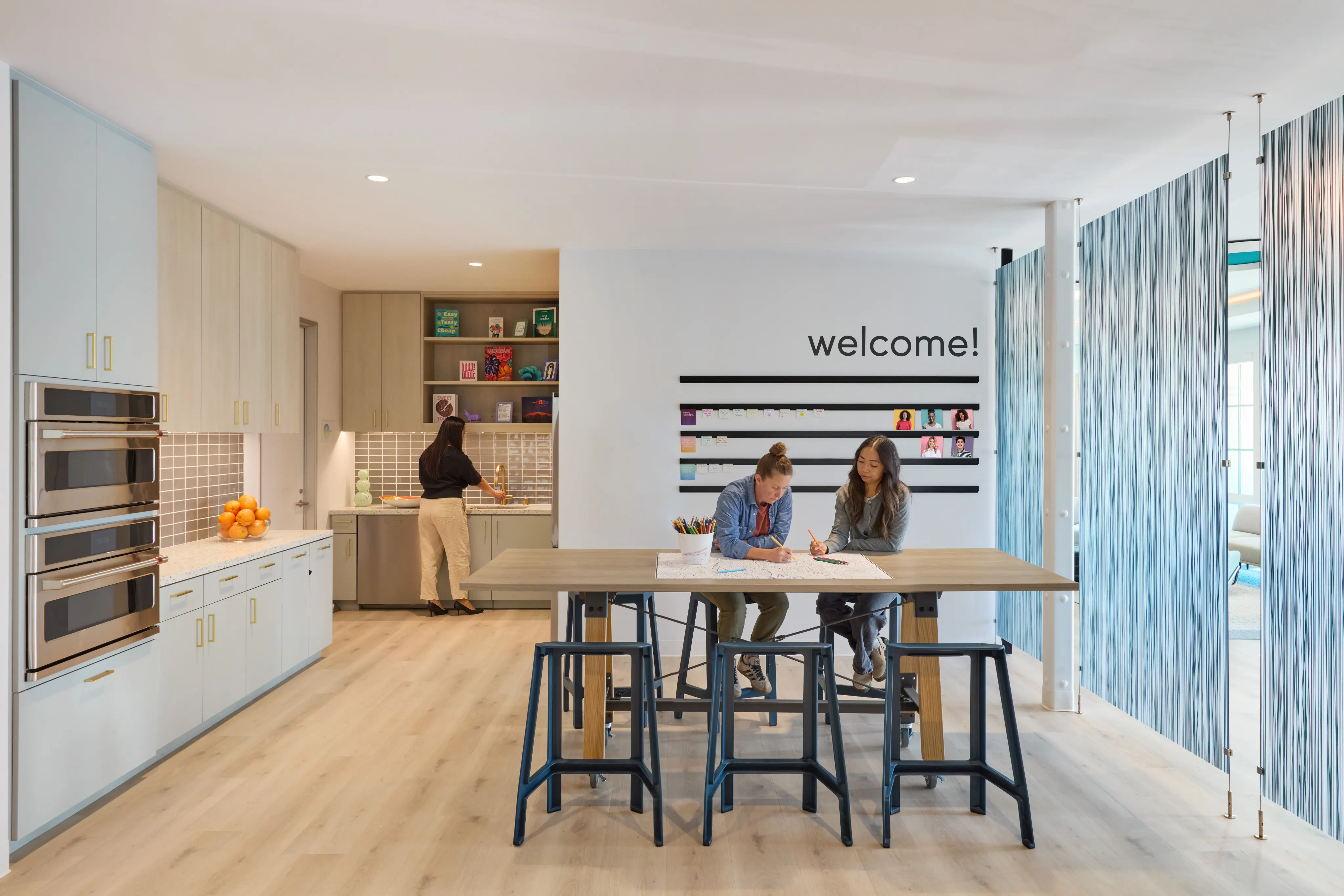 Modern room with a light wood floor and a kitchen on the left, featuring ovens and a counter with fruit. To the right, a central table with three stools has two people seated and writing. A welcoming ambiance is highlighted by a wall with "welcome!" and co